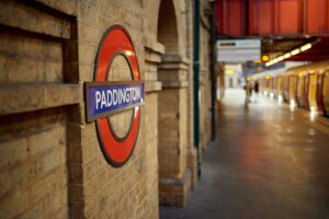 Insegna rotonda rossa e blu della metropolitana di Londra con la scritta 'PADDINGTON' su muro di mattoni color sabbia, con banchina sfocata sullo sfondo